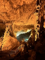 An underground cave scene exhibits striking rock formations with stalactites and stalagmites illuminated by warm lighting. A wooden walkway with railings leads through the cave, providing a path for exploration. The overall setting creates an enchanting and mysterious atmosphere.