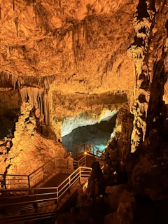 An underground cave scene exhibits striking rock formations with stalactites and stalagmites illuminated by warm lighting. A wooden walkway with railings leads through the cave, providing a path for exploration. The overall setting creates an enchanting and mysterious atmosphere.