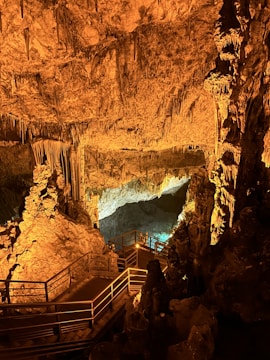An underground cave scene exhibits striking rock formations with stalactites and stalagmites illuminated by warm lighting. A wooden walkway with railings leads through the cave, providing a path for exploration. The overall setting creates an enchanting and mysterious atmosphere.