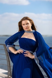 A woman stands confidently outdoors with her hands on her hips, wearing a flowing, elegant blue dress adorned with intricate patterns along the neckline and cuffs. The background features a blurred view of a body of water and a clear sky.