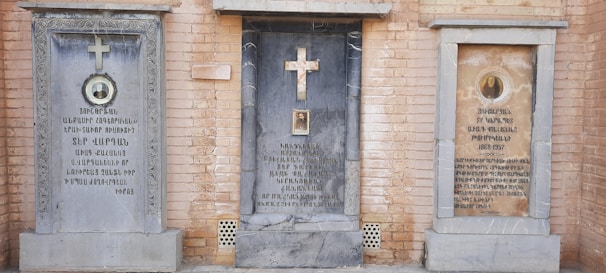 Three tombstones set against a brick wall, each with distinctive designs and inscriptions. The center tombstone features a prominent cross and inscriptions in a language with unique script. The left and right tombstones also have crosses and inscriptions, with additional circular portraits or medallions.