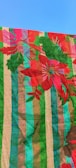 Colorful textile pattern featuring large red flowers and green leaves over a striped background. The stripes are composed of bold colors including green, blue, pink, and brown, creating a vibrant and striking design. The fabric is shown against a clear blue sky.