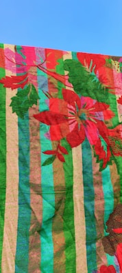 Colorful textile pattern featuring large red flowers and green leaves over a striped background. The stripes are composed of bold colors including green, blue, pink, and brown, creating a vibrant and striking design. The fabric is shown against a clear blue sky.