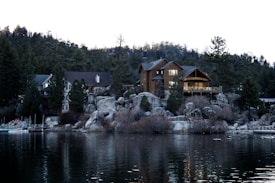 A serene lakeside scene featuring a large wooden cabin nestled among boulders and evergreen trees. The cabin is well-lit, suggesting warmth and coziness. The calm lake in the foreground reflects the structure and surrounding landscape, creating a peaceful atmosphere.
