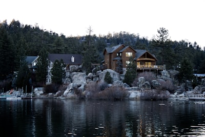 A serene lakeside scene featuring a large wooden cabin nestled among boulders and evergreen trees. The cabin is well-lit, suggesting warmth and coziness. The calm lake in the foreground reflects the structure and surrounding landscape, creating a peaceful atmosphere.