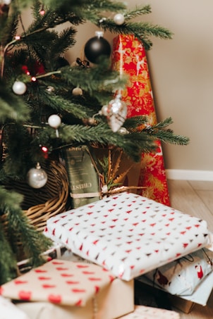A decorated Christmas tree with ornaments such as silver and black baubles. Several wrapped gifts in colorful paper featuring red and white patterns are placed underneath the tree. Pine needles and a red and gold fabric create a festive ambiance.
