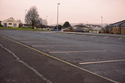 An empty parking lot with marked parking spaces and a few scattered leafless trees. A community center is visible to the left, and a row of residential houses stretches into the distance. A lone car is parked on the side of the road, and the scene is overcast, suggesting a cool or early morning atmosphere.