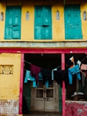 A vibrant yellow building with green shuttered windows on the upper level. Below, clothes of various colors, including blue jeans and black, red, and orange garments, hang on a line strung across an open area with two pink posts. The doorway is white and weathered, and the walls show signs of age.