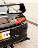 A glossy black sports car with a prominent rear spoiler is parked on a light-colored stone or brick surface. The car's taillights are illuminated with a distinctive red and amber glow, and the 'Supra Turbo' logo is visible on the back along with the Toyota emblem. The license plate is yellow and black with Arabic lettering.
