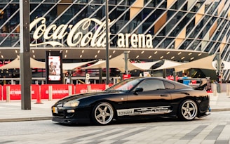 A sleek black sports car is parked in front of a modern arena with large glass panels and a prominent Coca-Cola sign. The car has customized decals and alloy wheels, and the scene is set in an urban environment with red banners and pavement.
