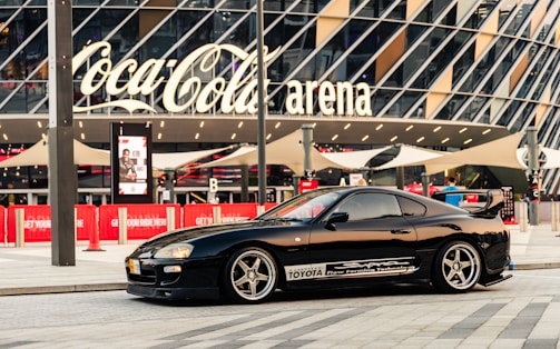 A sleek black sports car is parked in front of a modern arena with large glass panels and a prominent Coca-Cola sign. The car has customized decals and alloy wheels, and the scene is set in an urban environment with red banners and pavement.