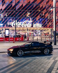 A sleek black sports car is parked in front of an illuminated arena entrance, with a large sign bearing the name of a famous beverage brand. The building features a modern, geometric facade with bright red and blue lights, creating a vibrant and lively atmosphere.