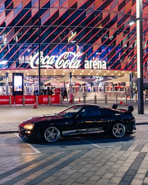 A sleek black sports car is parked in front of an illuminated arena entrance, with a large sign bearing the name of a famous beverage brand. The building features a modern, geometric facade with bright red and blue lights, creating a vibrant and lively atmosphere.