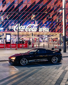 A sleek black sports car is parked in front of an illuminated arena entrance, with a large sign bearing the name of a famous beverage brand. The building features a modern, geometric facade with bright red and blue lights, creating a vibrant and lively atmosphere.