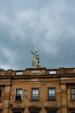 A stone statue of a robed figure holding a branch is positioned on the roof of a historic building with the word 'Italia' inscribed on its facade. The sky is overcast, casting a moody backdrop to the architecture.