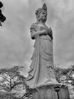 A large, intricately detailed statue portrays a figure wearing traditional attire and a headdress. The statue is surrounded by trees, and the sky overhead is cloudy, giving a dramatic backdrop.