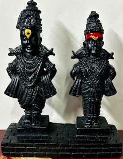 Two black statues with intricate details and headdresses stand side by side on a textured black base. Each statue displays rich ornamentation with multiple layers of jewelry and headgear. The left statue features a yellow marking on the forehead, while the right one has a red band and yellow marking.