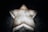A glowing, five-pointed star shape is set against a dark background. The star appears to be made of a material resembling frosted glass or ice, with a soft, diffuse glow emanating from it. Shadows and textures are visible on the surface around the star, emphasizing its three-dimensional structure.