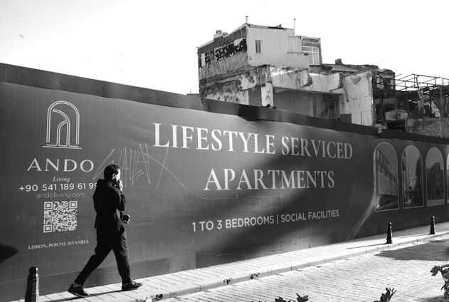 A person walks past a large advertising billboard for lifestyle serviced apartments. The billboard features text promoting one to three bedroom units with social facilities. In the background, a partially demolished or deteriorating building is visible. The scene is in black and white, adding a stark contrast to the modern advertisement and the old structure.