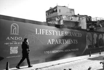 A person walks past a large advertising billboard for lifestyle serviced apartments. The billboard features text promoting one to three bedroom units with social facilities. In the background, a partially demolished or deteriorating building is visible. The scene is in black and white, adding a stark contrast to the modern advertisement and the old structure.