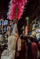 A street vendor carries bright pink cotton candy in transparent bags attached to long sticks. The scene is set in a busy, colorful market street at night, with various people walking and vibrant lights illuminating the area.