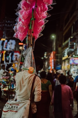 A street vendor carries bright pink cotton candy in transparent bags attached to long sticks. The scene is set in a busy, colorful market street at night, with various people walking and vibrant lights illuminating the area.