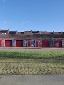 A row of garage doors painted in red with graffiti art on them, featuring stylized hearts and abstract designs. The building has a sloped roof and is set against a clear blue sky with some scattered clouds. In the foreground, there is a grassy area with a grid pattern visible on the ground.