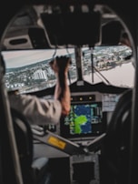 A view from inside an aircraft cockpit where a pilot is operating the controls. The landscape outside shows a city with numerous buildings and water bodies. A navigation screen is visible, providing flight information.