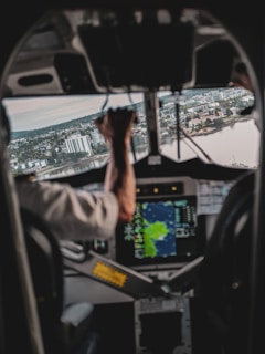 A view from inside an aircraft cockpit where a pilot is operating the controls. The landscape outside shows a city with numerous buildings and water bodies. A navigation screen is visible, providing flight information.
