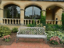 A beautifully landscaped garden features a wooden bench in the foreground, surrounded by lush greenery and colorful flowers. The backdrop consists of a stone building with elegant arched windows and ivy climbing the walls. There are ornamental balustrades and a brick path enhancing the tranquil setting.