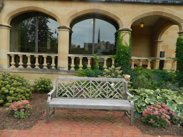 A beautifully landscaped garden features a wooden bench in the foreground, surrounded by lush greenery and colorful flowers. The backdrop consists of a stone building with elegant arched windows and ivy climbing the walls. There are ornamental balustrades and a brick path enhancing the tranquil setting.
