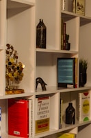 A neatly arranged bookshelf featuring a variety of books, some of which are titled 'Good to Great' and 'The 7 Habits of Highly Effective People'. A decorative metal sculpture resembling a tree with round, golden leaves sits prominently on the shelf, along with marble-patterned vases. Small potted plants add a touch of greenery, while a bicycle-themed book provides a whimsical touch.