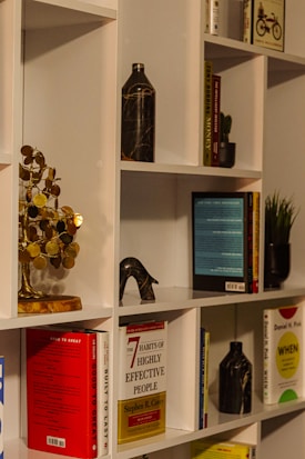 A neatly arranged bookshelf featuring a variety of books, some of which are titled 'Good to Great' and 'The 7 Habits of Highly Effective People'. A decorative metal sculpture resembling a tree with round, golden leaves sits prominently on the shelf, along with marble-patterned vases. Small potted plants add a touch of greenery, while a bicycle-themed book provides a whimsical touch.