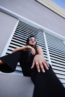A woman sits confidently, looking upwards while wearing contemporary sunglasses. The perspective is from a low angle, accentuating her pose. She's dressed in dark, fashionable clothing and is positioned against a background of horizontal metal slats, likely part of a modern architectural structure.