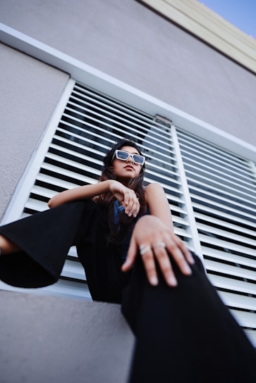 A woman sits confidently, looking upwards while wearing contemporary sunglasses. The perspective is from a low angle, accentuating her pose. She's dressed in dark, fashionable clothing and is positioned against a background of horizontal metal slats, likely part of a modern architectural structure.