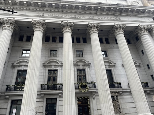 An ornate building facade with large, classical columns and a sign reading 'The Canadian Bank of Commerce.' The architecture features detailed stonework and decorative wreaths hanging on the railings.