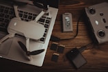 A drone with propellers is placed on a laptop keyboard alongside a battery, a remote controller, multiple SD cards, and a memory card reader on a wooden surface. The arrangement of these items suggests preparation for a video or photography session.