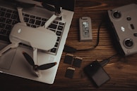 A drone with propellers is placed on a laptop keyboard alongside a battery, a remote controller, multiple SD cards, and a memory card reader on a wooden surface. The arrangement of these items suggests preparation for a video or photography session.