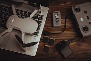 A drone with propellers is placed on a laptop keyboard alongside a battery, a remote controller, multiple SD cards, and a memory card reader on a wooden surface. The arrangement of these items suggests preparation for a video or photography session.