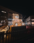 A large teddy bear figure illuminated with numerous small lights stands prominently in a dark urban setting. Nearby, a decorative gift box is also adorned with lights. The scene captures a festive display, likely in a public area, with buildings and evening sky in the background.