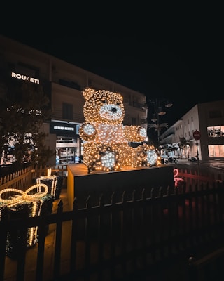 A large teddy bear figure illuminated with numerous small lights stands prominently in a dark urban setting. Nearby, a decorative gift box is also adorned with lights. The scene captures a festive display, likely in a public area, with buildings and evening sky in the background.