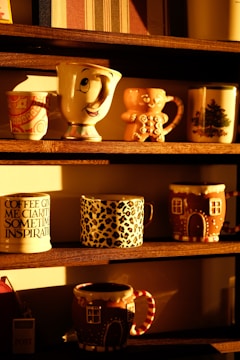 A collection of decorative mugs is arranged on wooden shelves, each with unique designs. Some mugs feature whimsical patterns, such as a mug with a smiling face, and others have seasonal or animal print designs. The sunlight casts a warm glow, enhancing the cozy and inviting atmosphere.