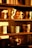A collection of decorative mugs is arranged on wooden shelves, each with unique designs. Some mugs feature whimsical patterns, such as a mug with a smiling face, and others have seasonal or animal print designs. The sunlight casts a warm glow, enhancing the cozy and inviting atmosphere.