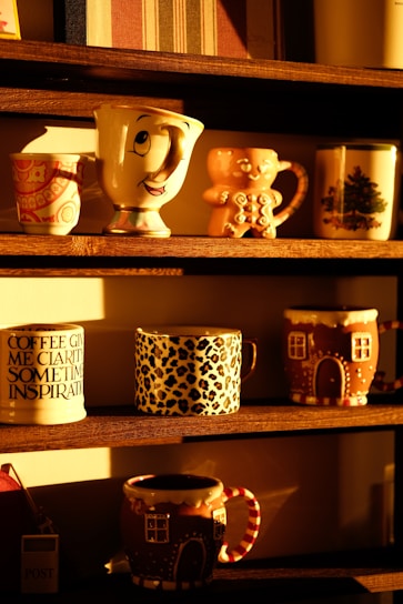 A collection of decorative mugs is arranged on wooden shelves, each with unique designs. Some mugs feature whimsical patterns, such as a mug with a smiling face, and others have seasonal or animal print designs. The sunlight casts a warm glow, enhancing the cozy and inviting atmosphere.