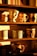 A collection of decorative mugs is arranged on wooden shelves, each with unique designs. Some mugs feature whimsical patterns, such as a mug with a smiling face, and others have seasonal or animal print designs. The sunlight casts a warm glow, enhancing the cozy and inviting atmosphere.