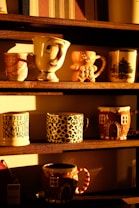 A collection of decorative mugs is arranged on wooden shelves, each with unique designs. Some mugs feature whimsical patterns, such as a mug with a smiling face, and others have seasonal or animal print designs. The sunlight casts a warm glow, enhancing the cozy and inviting atmosphere.