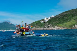 A boat filled with people is sailing on the sea near a hilly coastline. The boat appears festive with colorful decorations and passengers standing and sitting on the top deck. In the background, there are white buildings on the green hillside, possibly houses or resorts. The sky is clear with some clouds near the horizon.