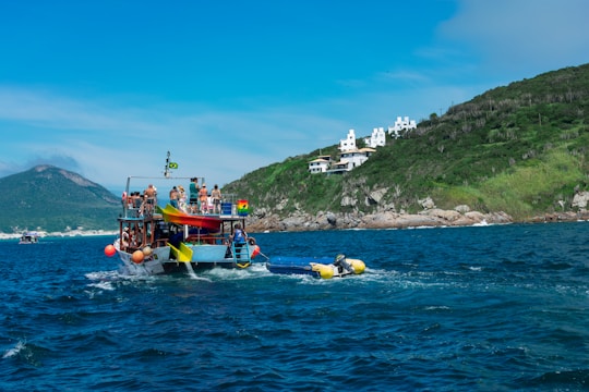 A boat filled with people is sailing on the sea near a hilly coastline. The boat appears festive with colorful decorations and passengers standing and sitting on the top deck. In the background, there are white buildings on the green hillside, possibly houses or resorts. The sky is clear with some clouds near the horizon.