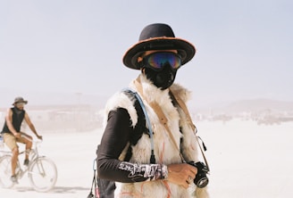 A person dressed in artistic attire stands in a desert setting, wearing a black hat with an orange rim, mirrored goggles, and a gas mask. Their outfit includes a furry vest and various accessories, including a camera they hold. In the background, another person rides a bicycle amidst a hazy atmosphere, suggesting a festival or event.
