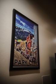 A framed artwork depicting a vibrant street scene in Park City. The illustration shows colorful buildings lining the street with mountains visible in the background. The cars are parked along the roadside, adding to the urban feel.